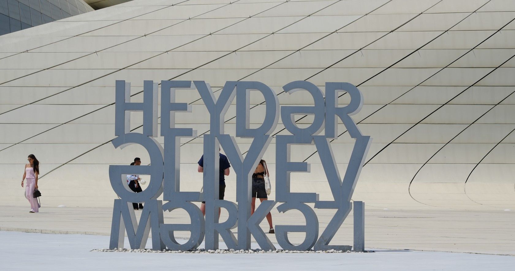 Groot bord met de tekst HEYDAR ALIYEV MƏRKƏZİ voor een wit, gebogen gebouw, met een persoon die er vlakbij loopt.