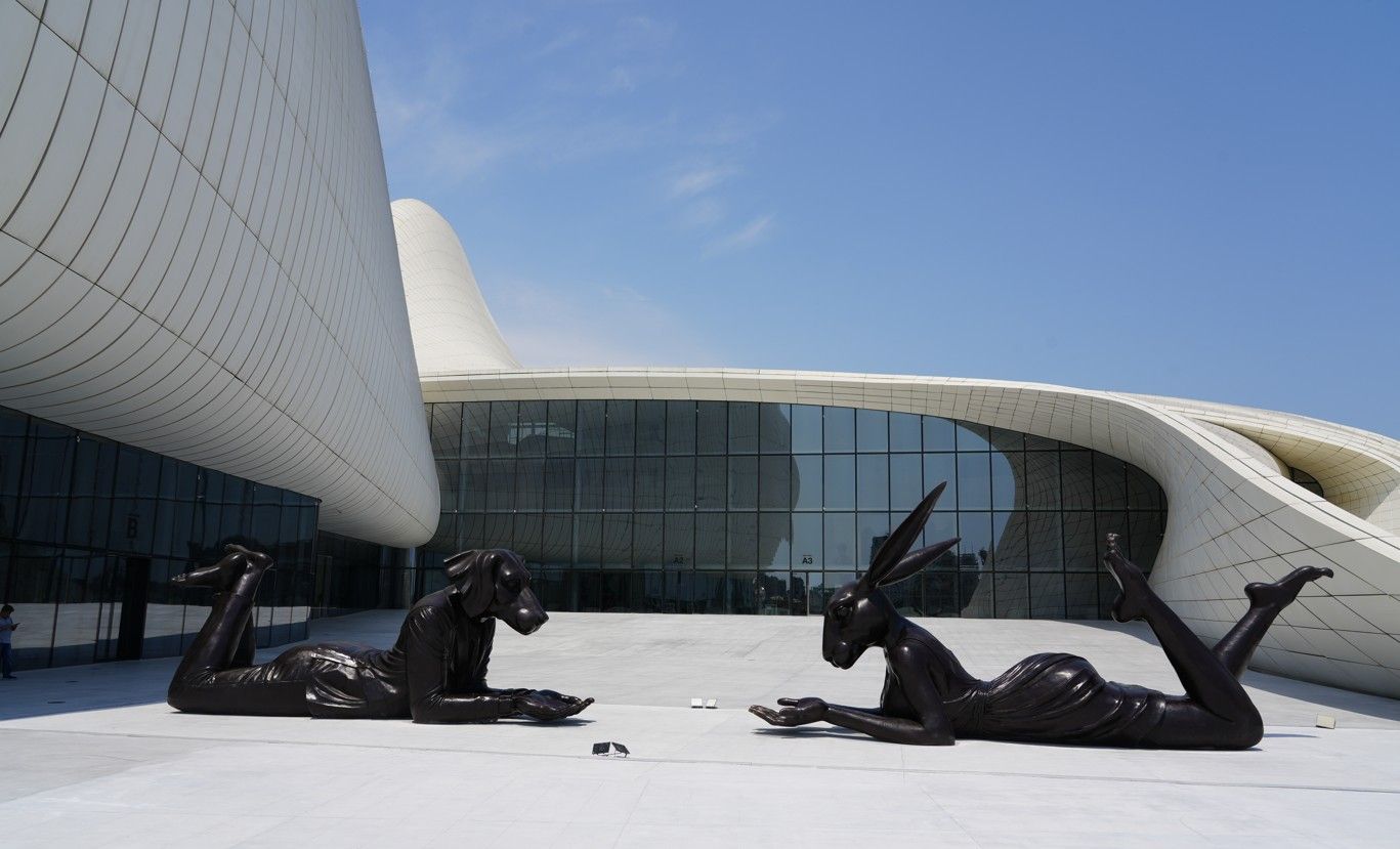 Modern wit gebouw met glazen gevel en donkere sculpturen op een plein onder een blauwe hemel.