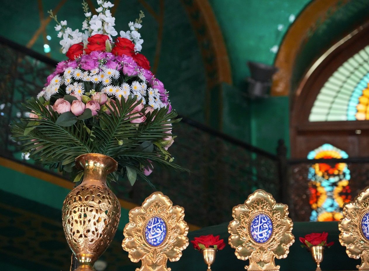 Een rijk versierd bloemstuk in een gouden vaas in een kerk, met glas-in-loodramen en kaarsen op de achtergrond.
