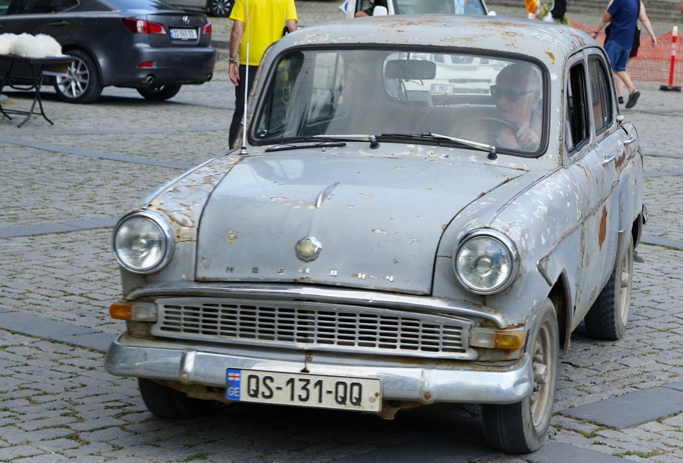 Een oude, grijze oldtimer staat geparkeerd op een straat met kinderkopjes, met mensen en andere auto's op de achtergrond.