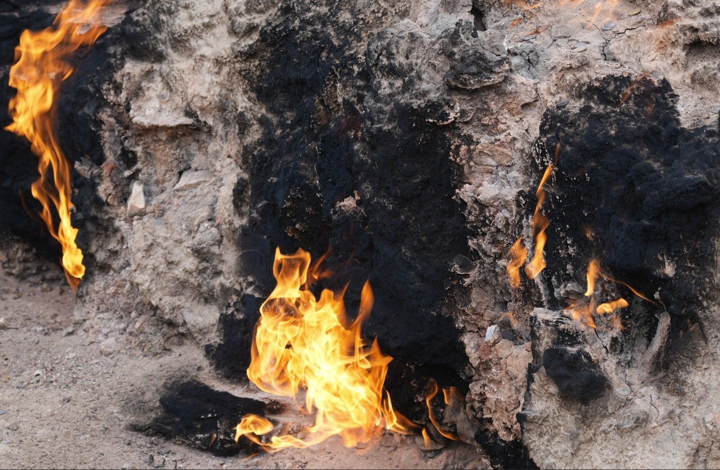 Verkoolde, brandende grond met vlammen die flikkeren tussen zwartgeblakerde rotsen en as.