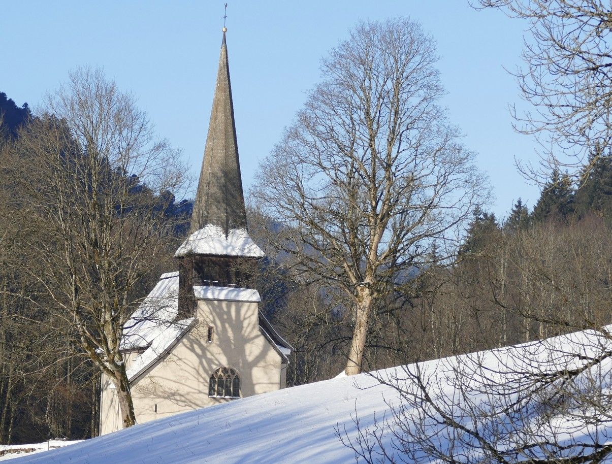 Een met sneeuw bedekte heuvel met een kleine kapel, een hoge torenspits en kale bomen onder een heldere, blauwe hemel.