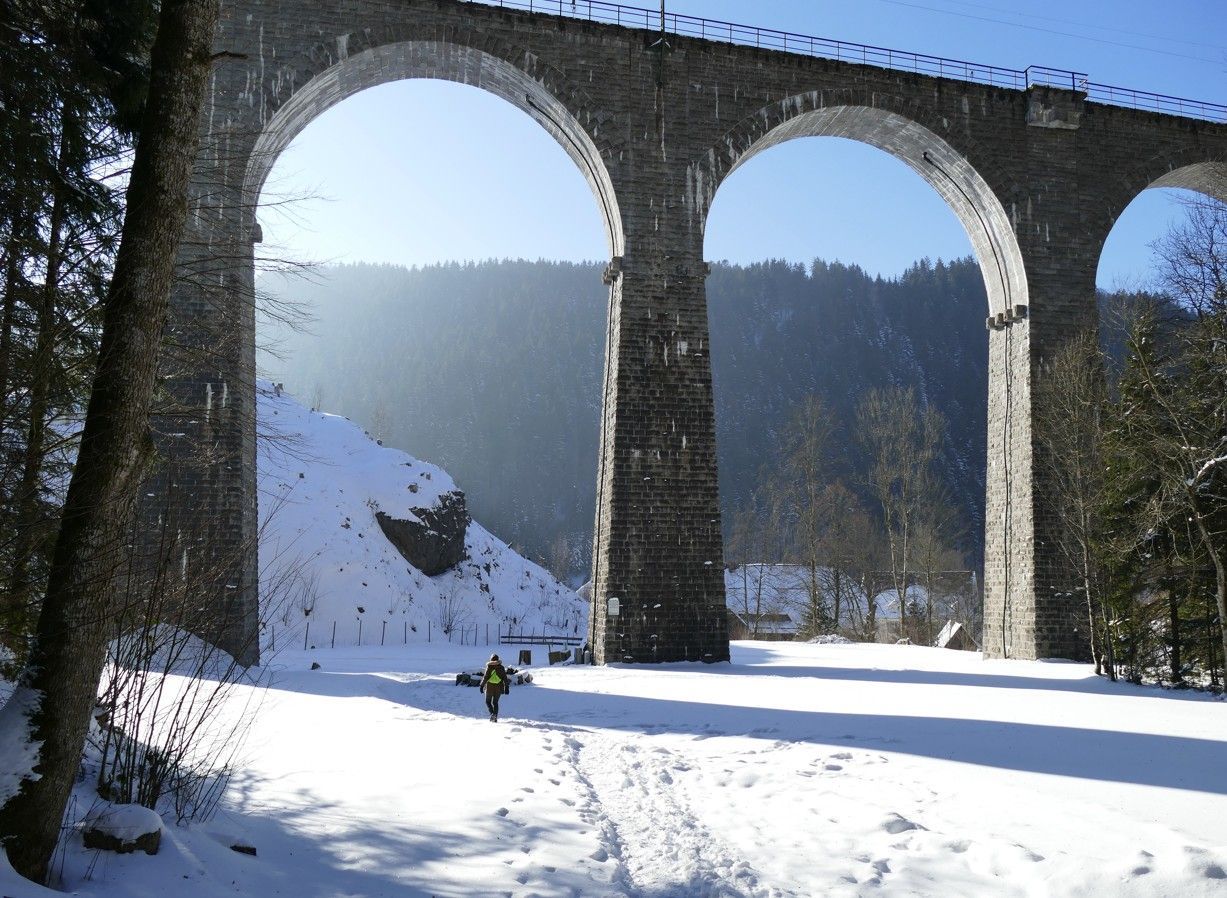Een besneeuwd tafereel onder een hoog stenen viaduct met bogen; iemand loopt over een besneeuwd pad richting de bergen.