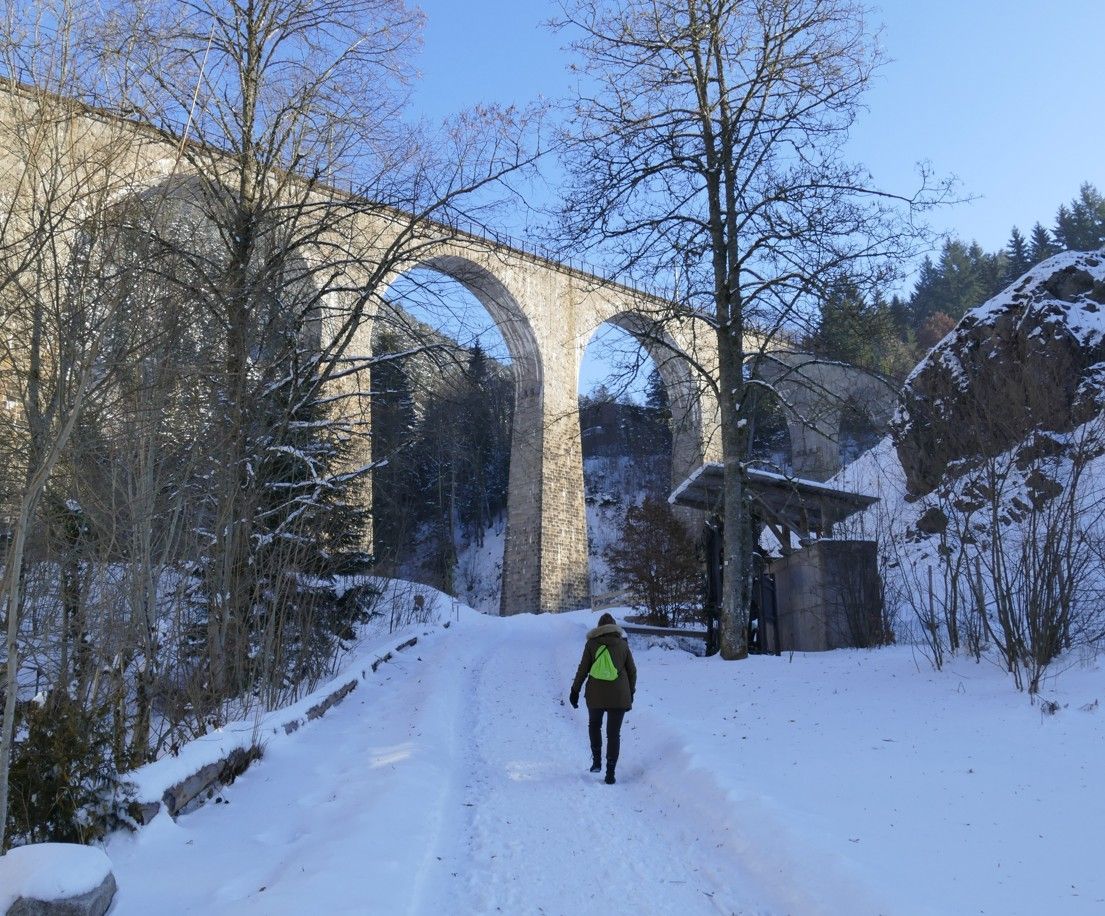 Een persoon loopt over een besneeuwd pad naar een grote stenen brug. Winterlandschap met bomen en een blauwe lucht.