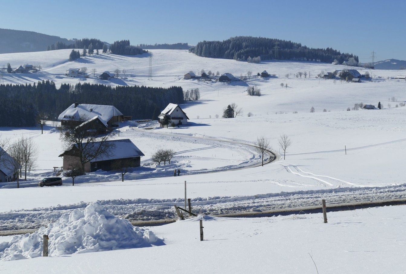 Een met sneeuw bedekt landschap met huizen en een kronkelende weg onder een helderblauwe hemel.