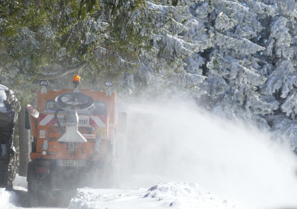 Een oranje sneeuwploeg sproeit sneeuw op een met sneeuw bedekte weg in de buurt van met sneeuw beladen bomen.