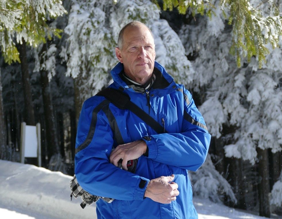 Een man in een blauwe jas staat in een besneeuwd bos, met op de achtergrond bomen die eveneens met sneeuw bedekt zijn.