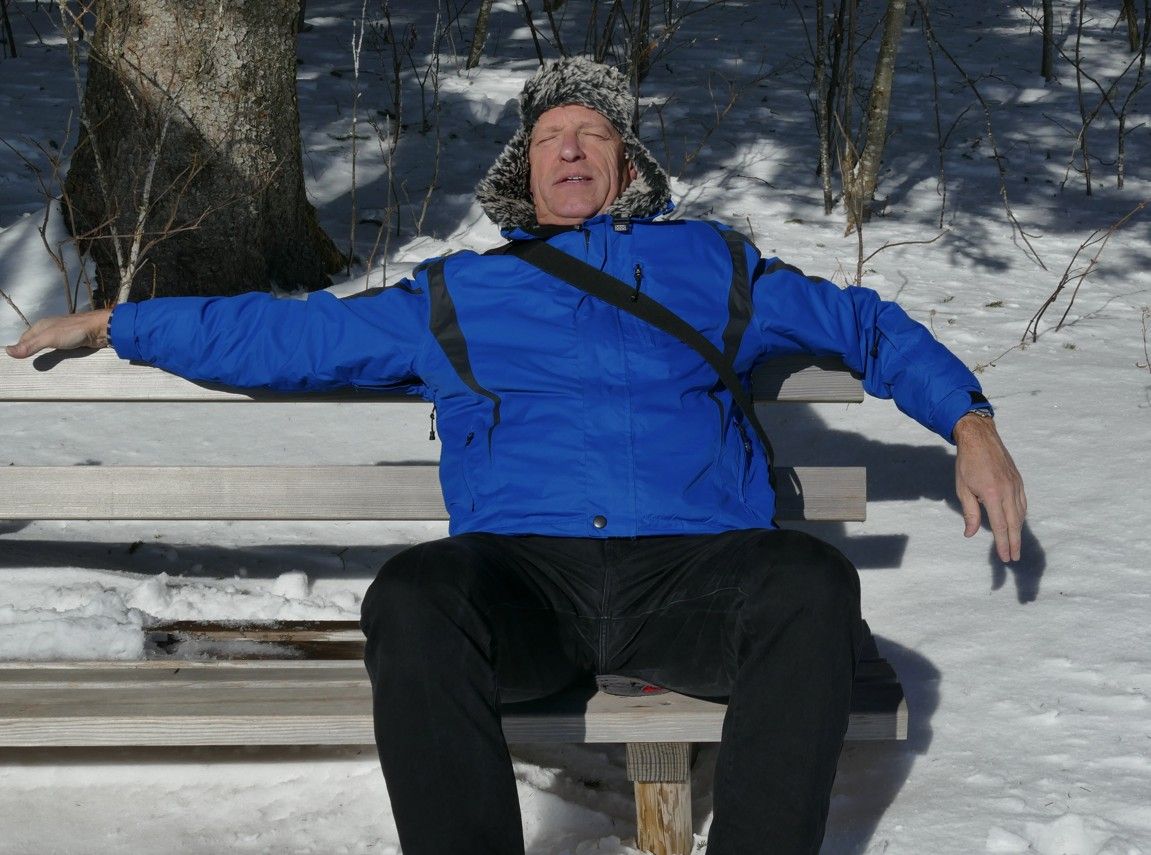 Een man in een blauwe jas en bontmuts zit met uitgestrekte armen op een besneeuwde parkbank.