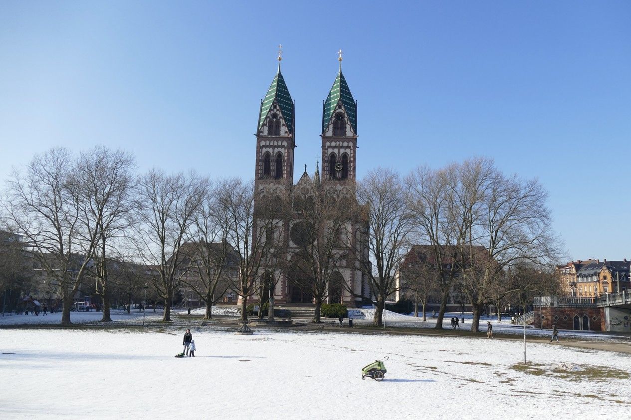 Een kerk met twee groene koepeltorens tegen een helderblauwe hemel, een besneeuwde voorgrond en kale bomen.