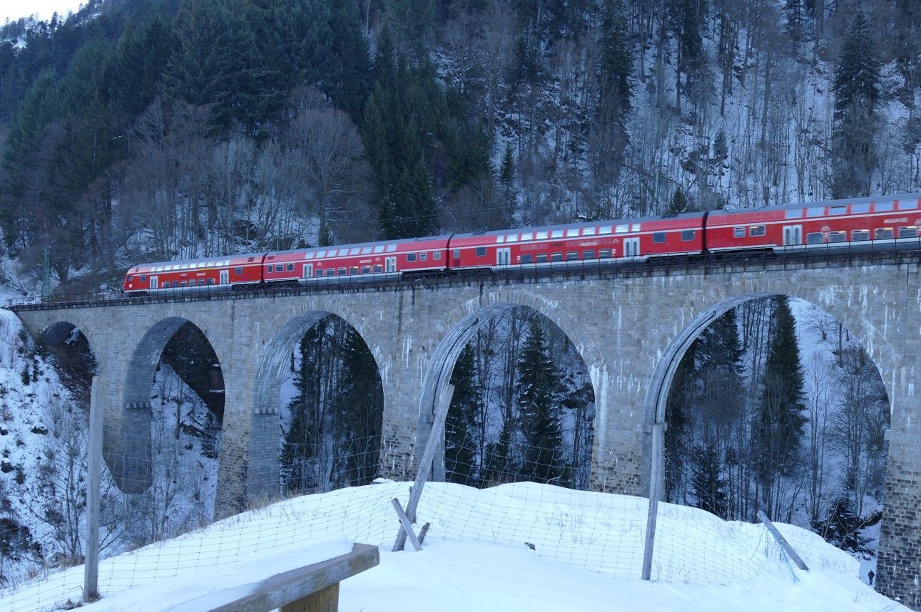 Een rode trein rijdt over een stenen boogbrug in een besneeuwd berglandschap.