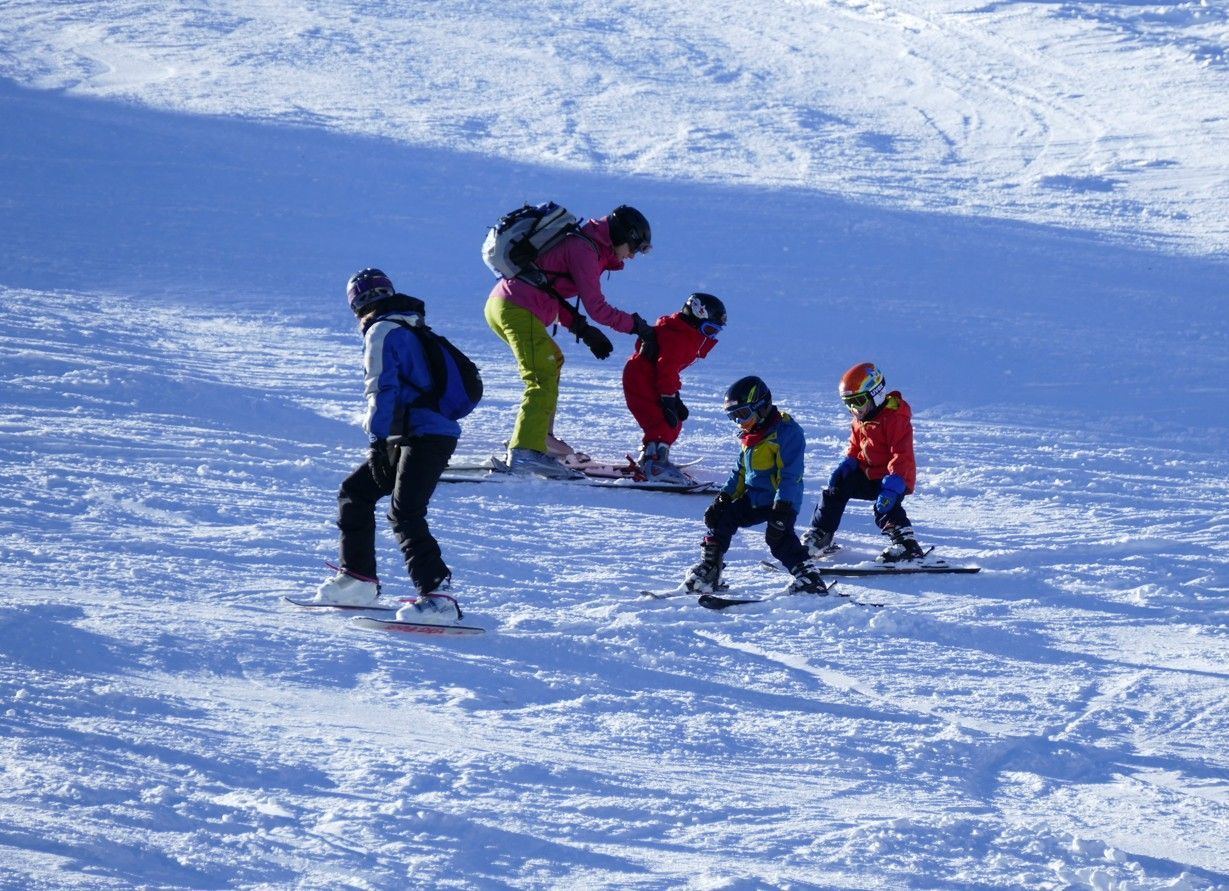 Een groep skiërs, een instructeur helpt een kind in het rood, anderen skiën op een besneeuwde helling.