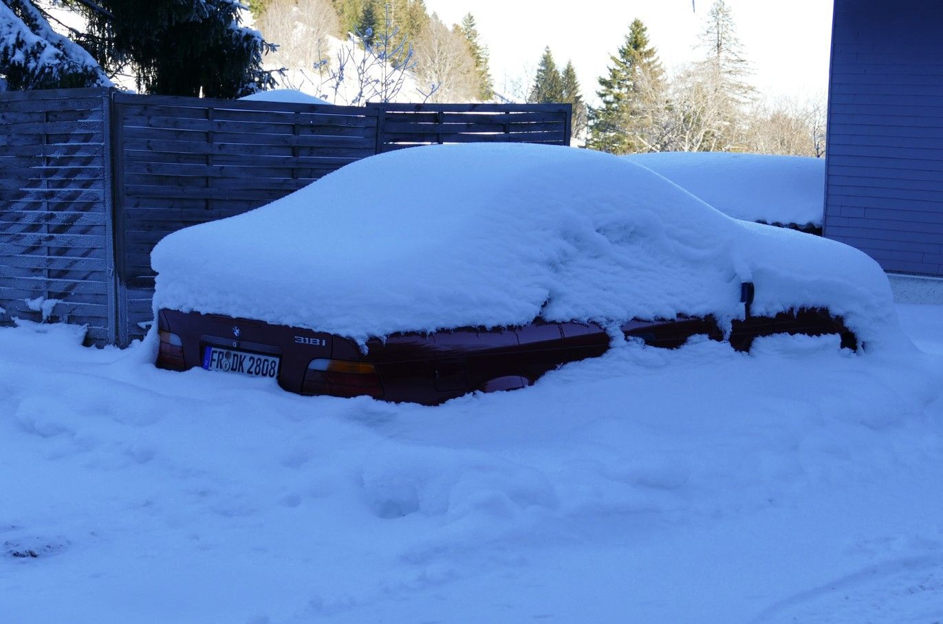 Een rode auto, volledig bedekt met sneeuw, staat geparkeerd buiten bij een houten hek en een gebouw op een zonnige winterdag.