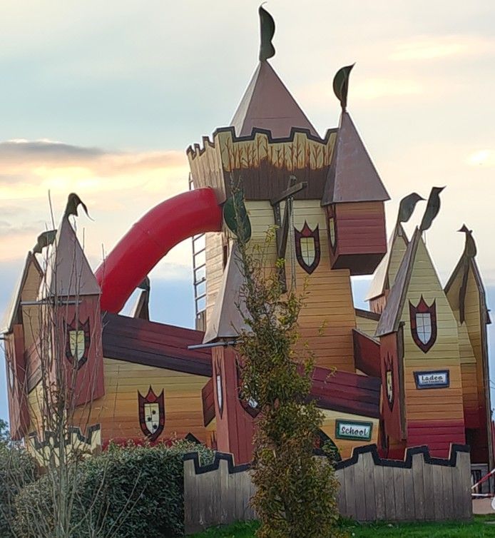 Speelkasteel met rode glijbaan, bruin en oranje geverfd hout, vlaggen en decoratieve schilden.