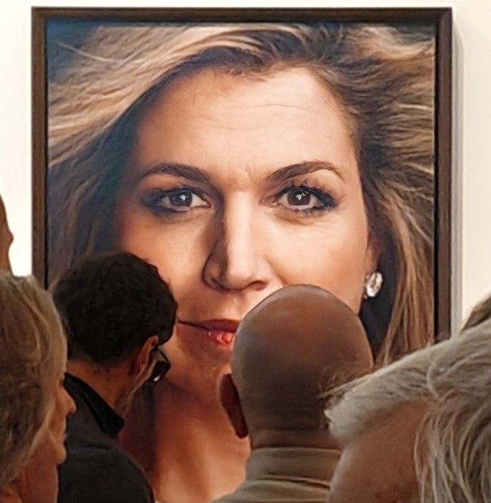 Mensen bekijken een groot portret van een vrouw met blond haar en oorbellen op een kunsttentoonstelling.