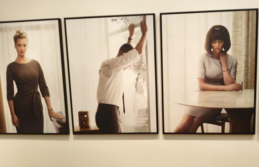 Aan de muur hangen drie ingelijste foto's: een vrouw in een jurk, een man die iets optilt en een vrouw aan een tafel.