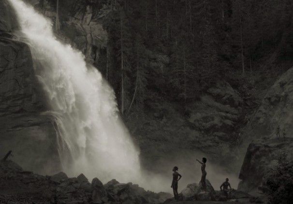 Waterval met vier figuren op rotsen. Omgeven door bos. Zwart-wit.