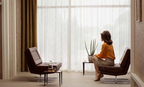 Vrouw zit en kijkt uit een groot raam; bruine stoelen, kleine tafel; lichtgekleurde rok, oranje top.