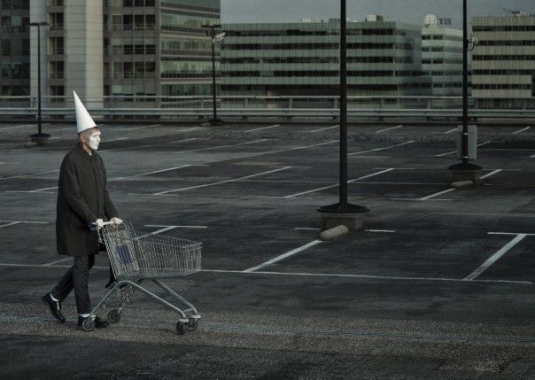 Een persoon met een witte puntmuts duwt een winkelwagen over een leeg parkeerterrein, met grijze gebouwen op de achtergrond.