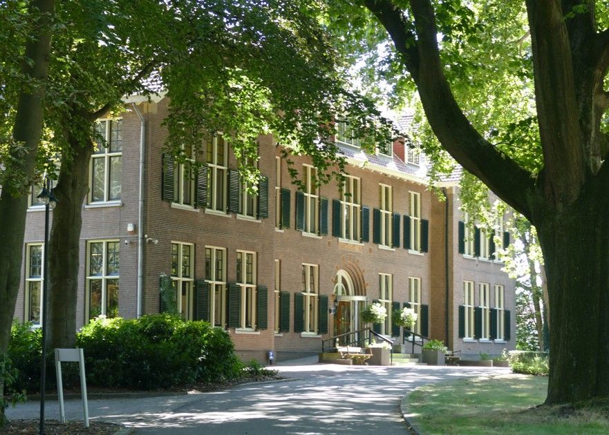 Een bakstenen gebouw met meerdere ramen, luiken en een schuin dak, omgeven door bomen.