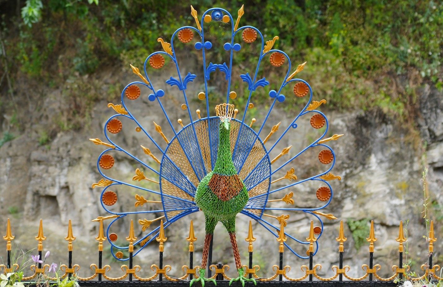 Een pauwvormig beeldhouwwerk met blauwe en oranje accenten in een tuin, met op de achtergrond een rotswand en groen.