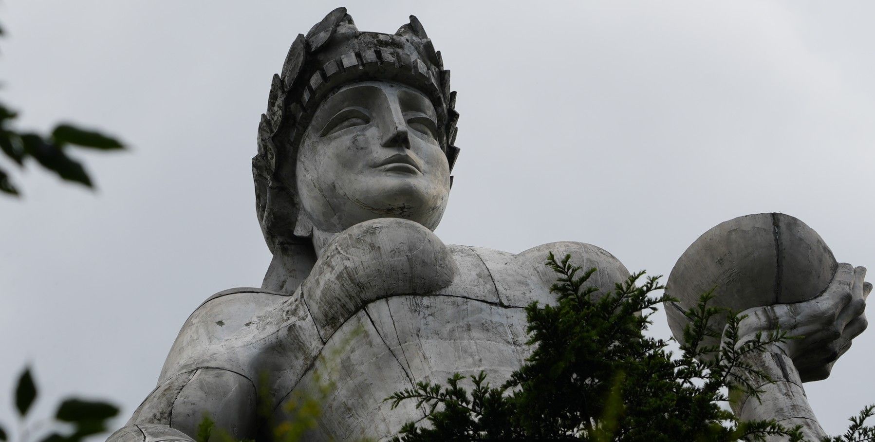 Groot stenen beeld van een gekroonde figuur, van onderaf gezien tegen een bewolkte hemel, met boomtakken op de voorgrond.