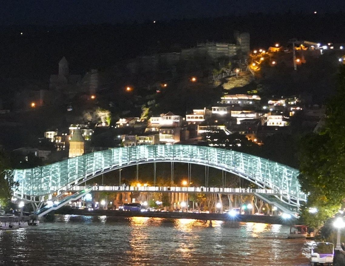 Nachtelijk uitzicht op een verlichte boogbrug over een rivier, met stadslichten en heuvels op de achtergrond.