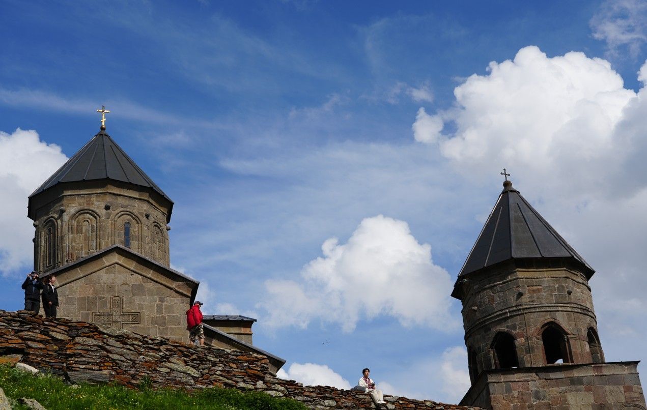 Stenen kerktorens met donkere daken tegen een blauwe hemel, met mensen op de stenen muur eronder.