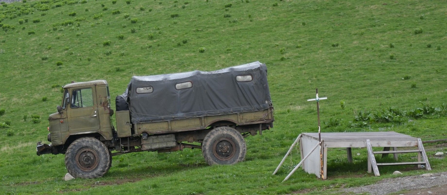 Een olijfgroene militaire vrachtwagen met afgedekte laadbak staat geparkeerd op een grashelling bij een kleine houten schuilplaats.