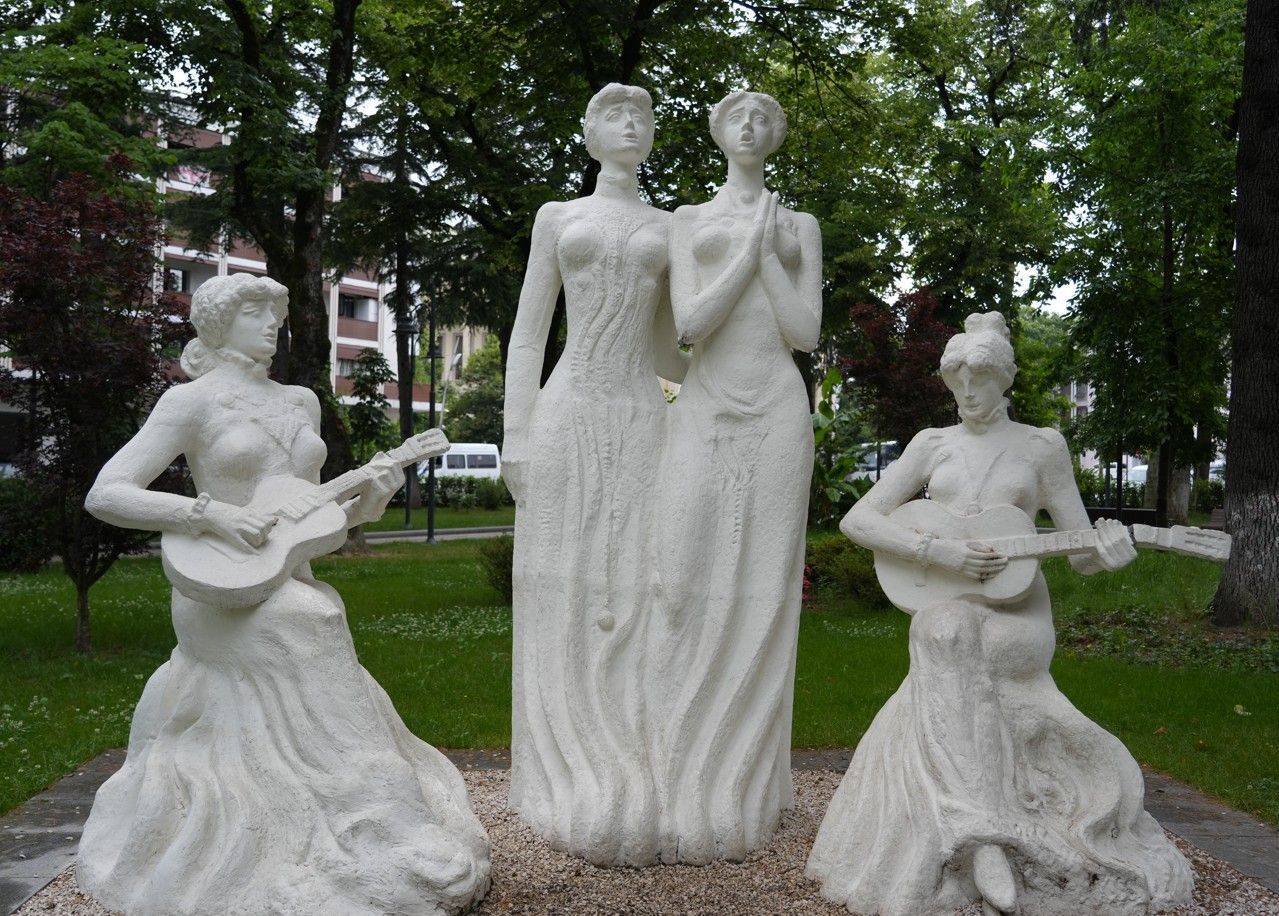Witte sculptuur van drie zittende figuren in een park, waarvan twee een gitaar vasthouden naast een lange centrale figuur.