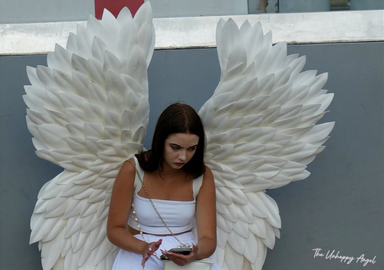 Vrouw in een witte jurk met grote witte engelenvleugels, die tegen een grijze muur naar haar telefoon kijkt.