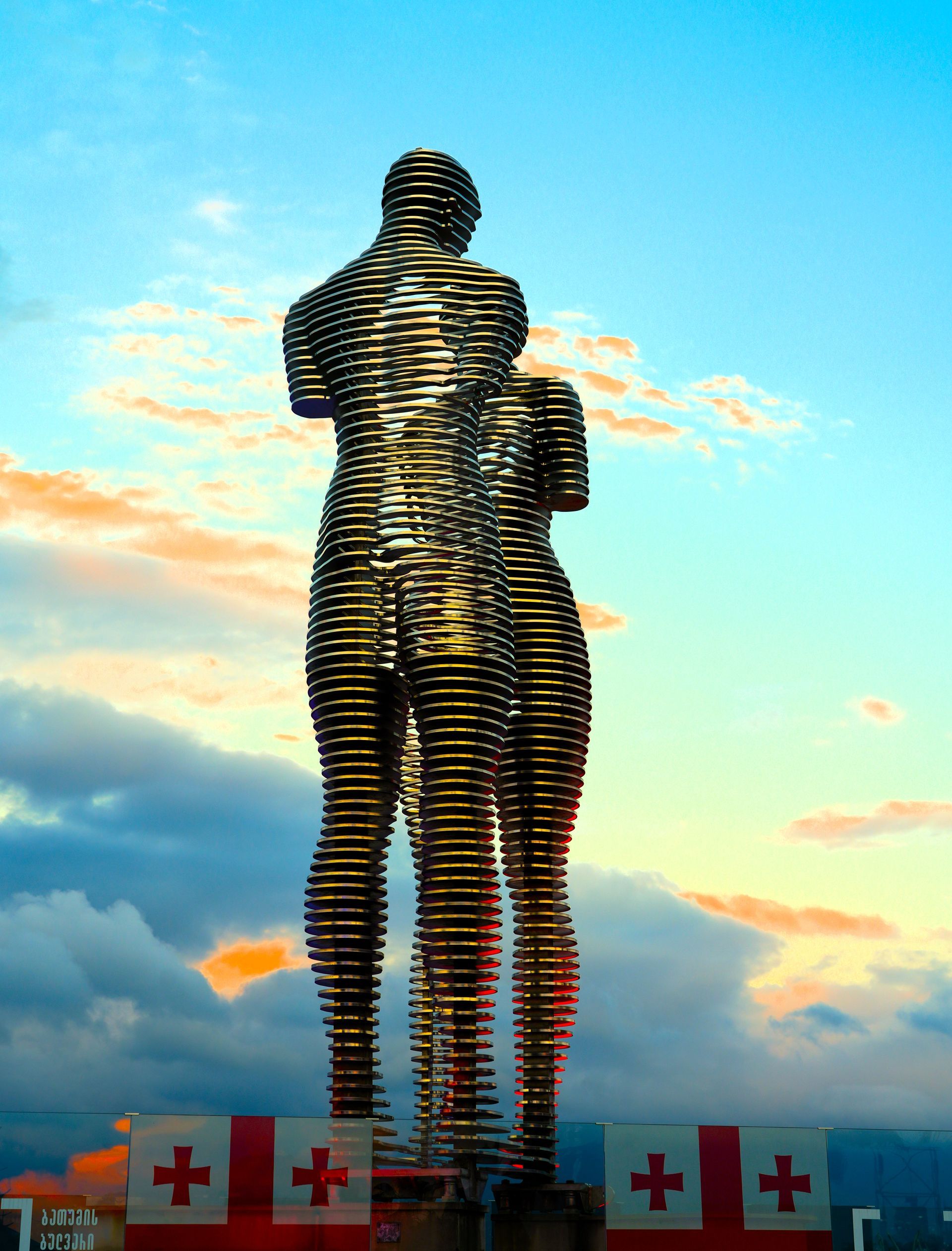 Opvallende verticale sculptuur tegen een zonsondergang, met rode kruisvlaggen langs de voet.