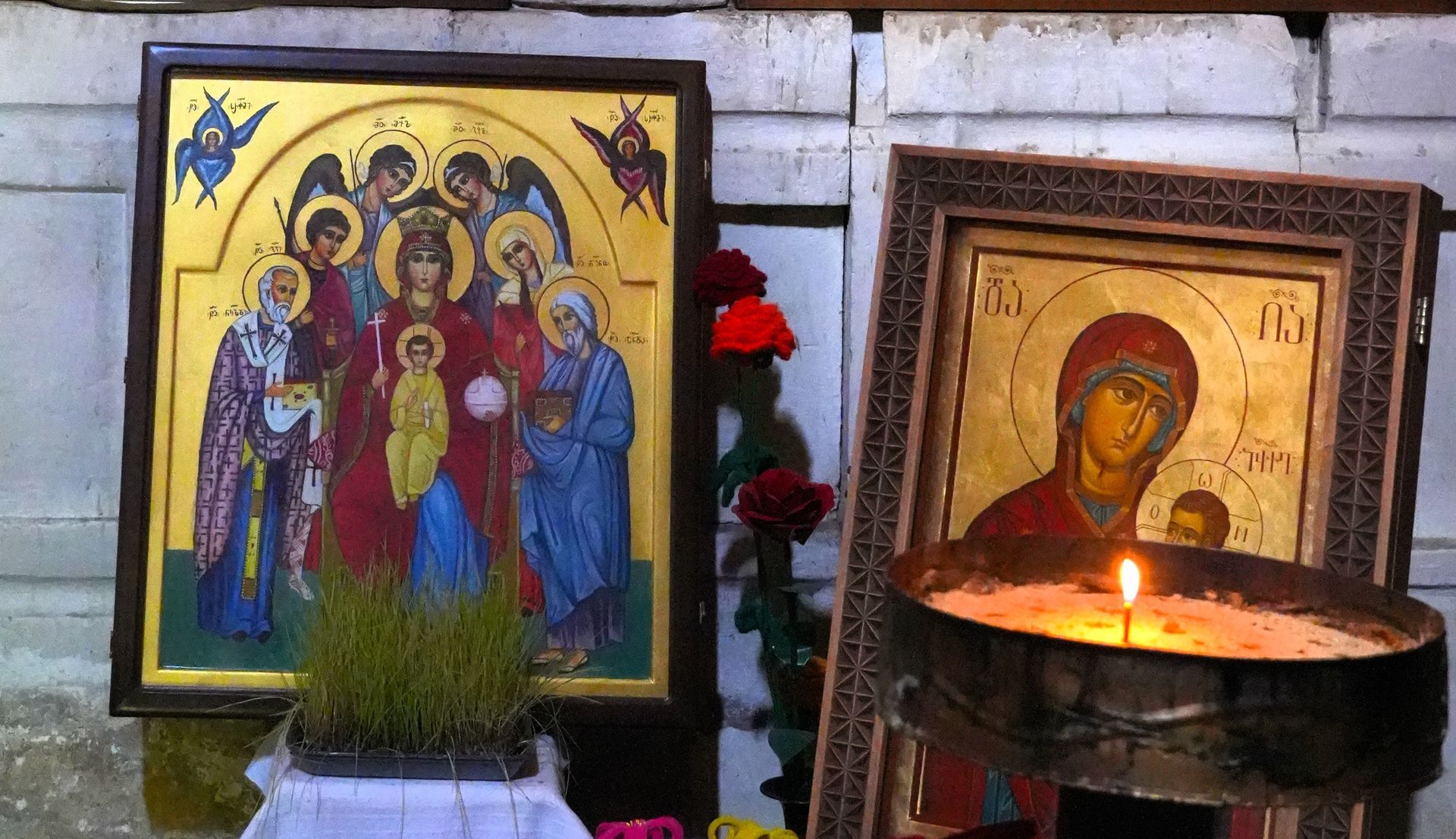 Twee ingelijste orthodoxe iconen met een brandende kaars ervoor, tegen een witte bakstenen muur.
