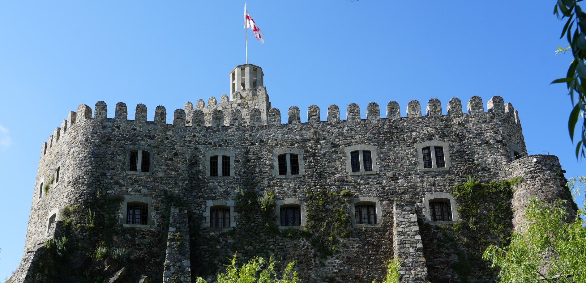 Stenen kasteel met kantelen en een wapperende vlag, gezien van onderaf tegen een helderblauwe hemel.