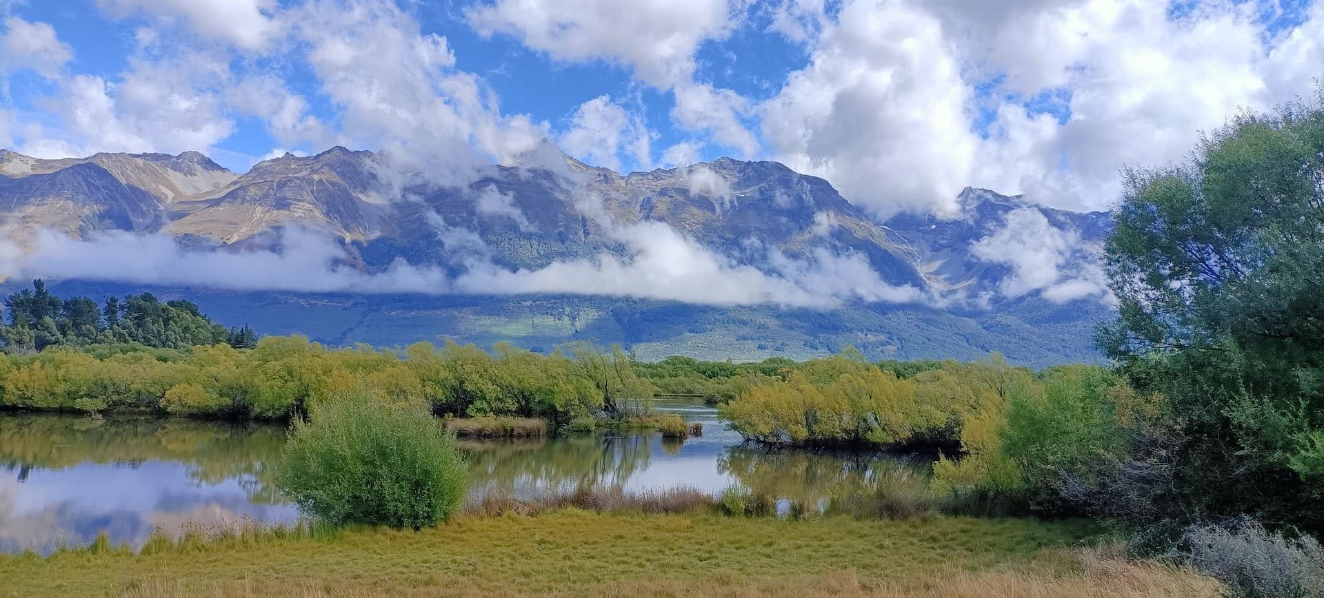 Bergen, gedeeltelijk bedekt met wolken, doemen op boven een kalm meer en groen gebladerte onder een blauwe hemel.