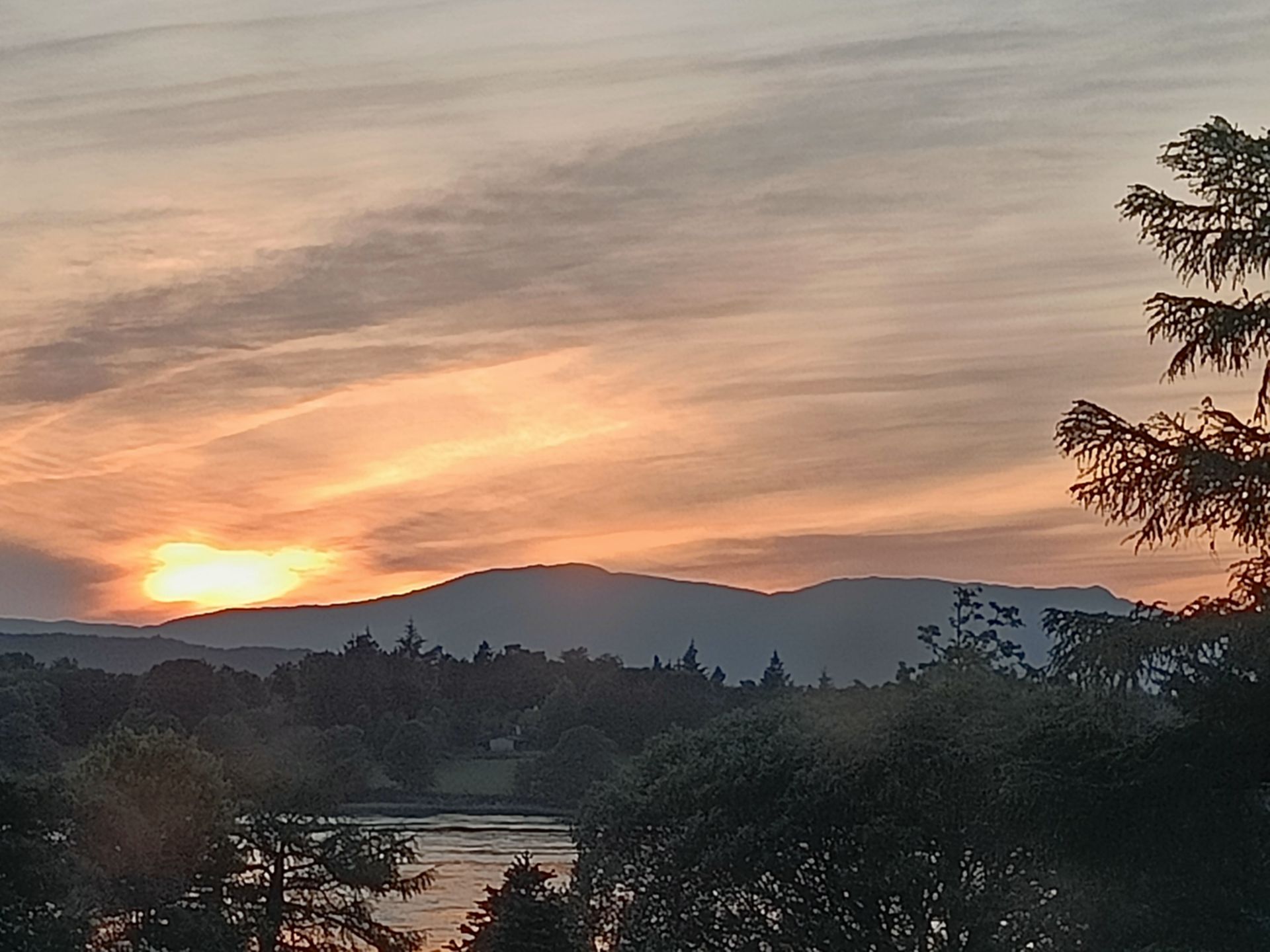 Zonsondergang boven silhouetten van bergen, bomen en water, met een oranje en perzikkleurige lucht.