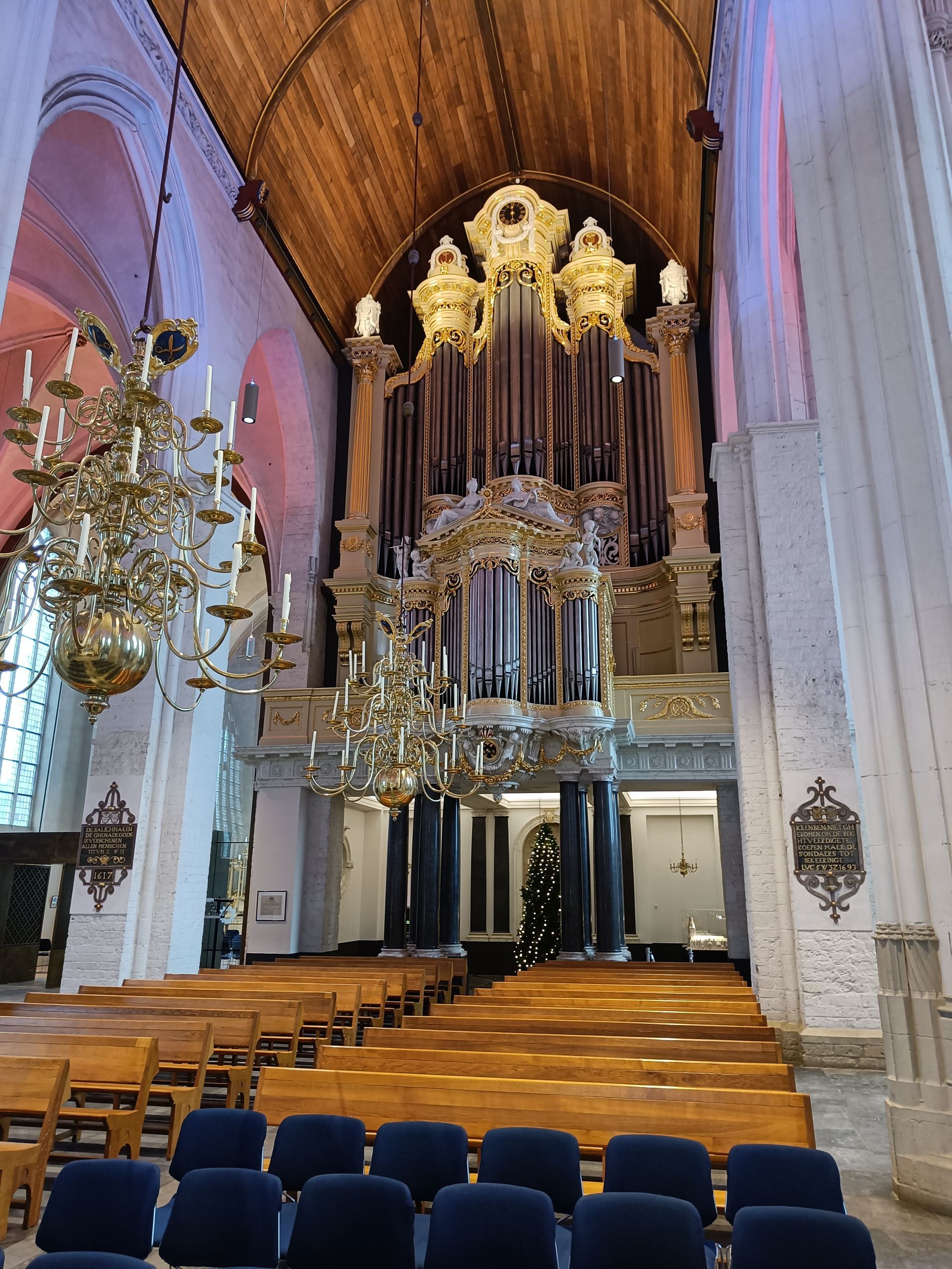 In een kerk domineert een groot pijporgel de achterwand boven rijen kerkbanken. Aan de linkerkant hangen kroonluchters.
