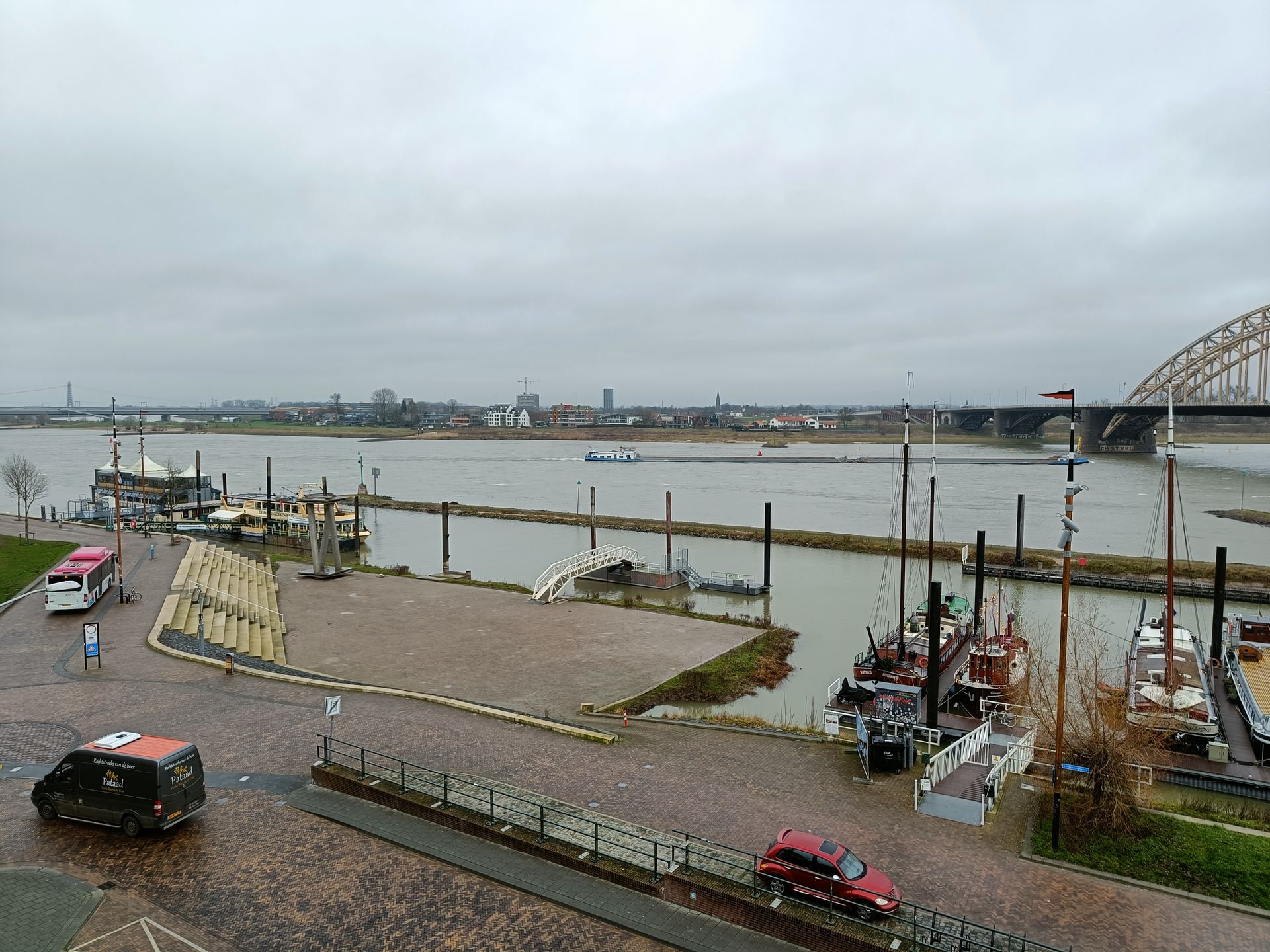 Uitzicht op een rivier met aangemeerde boten, een brug en een stadsgezicht onder een bewolkte hemel.