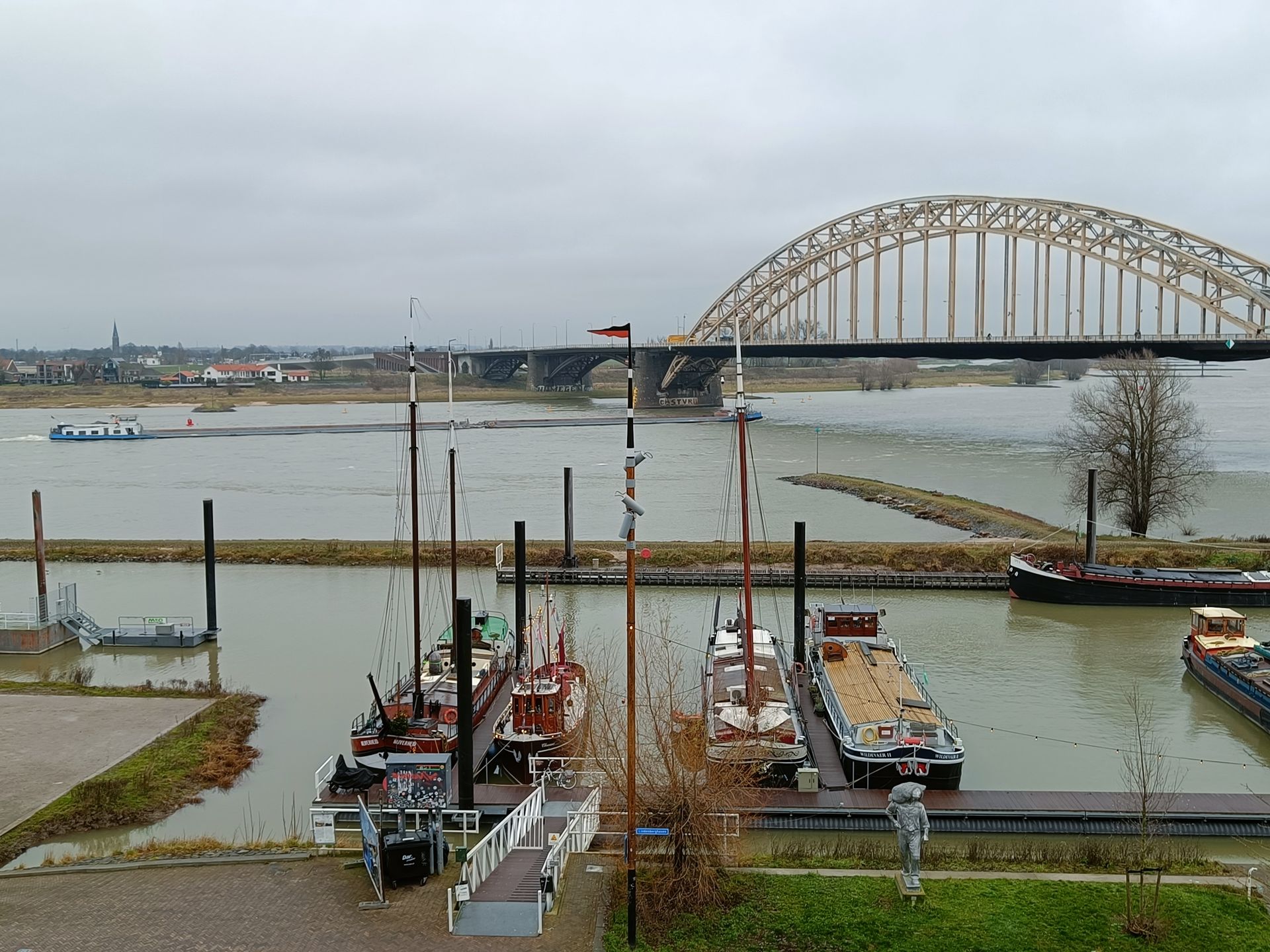 Boten aangemeerd in een haven vlakbij een brug, met de rivier op de achtergrond. Bewolkte lucht.
