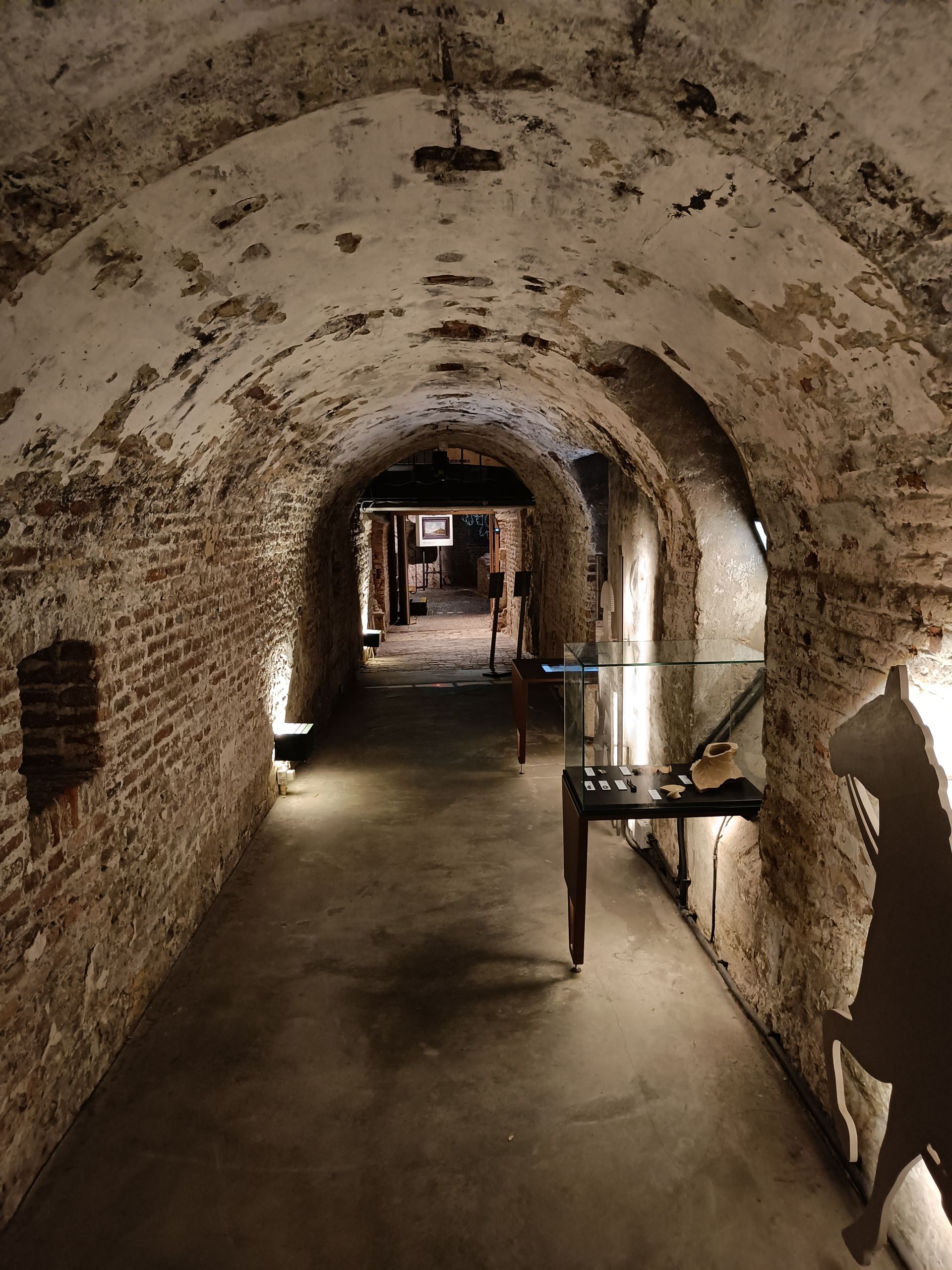 Bakstenen tunnel met gewelfd plafond, verlicht pad met vitrines en artefacten.