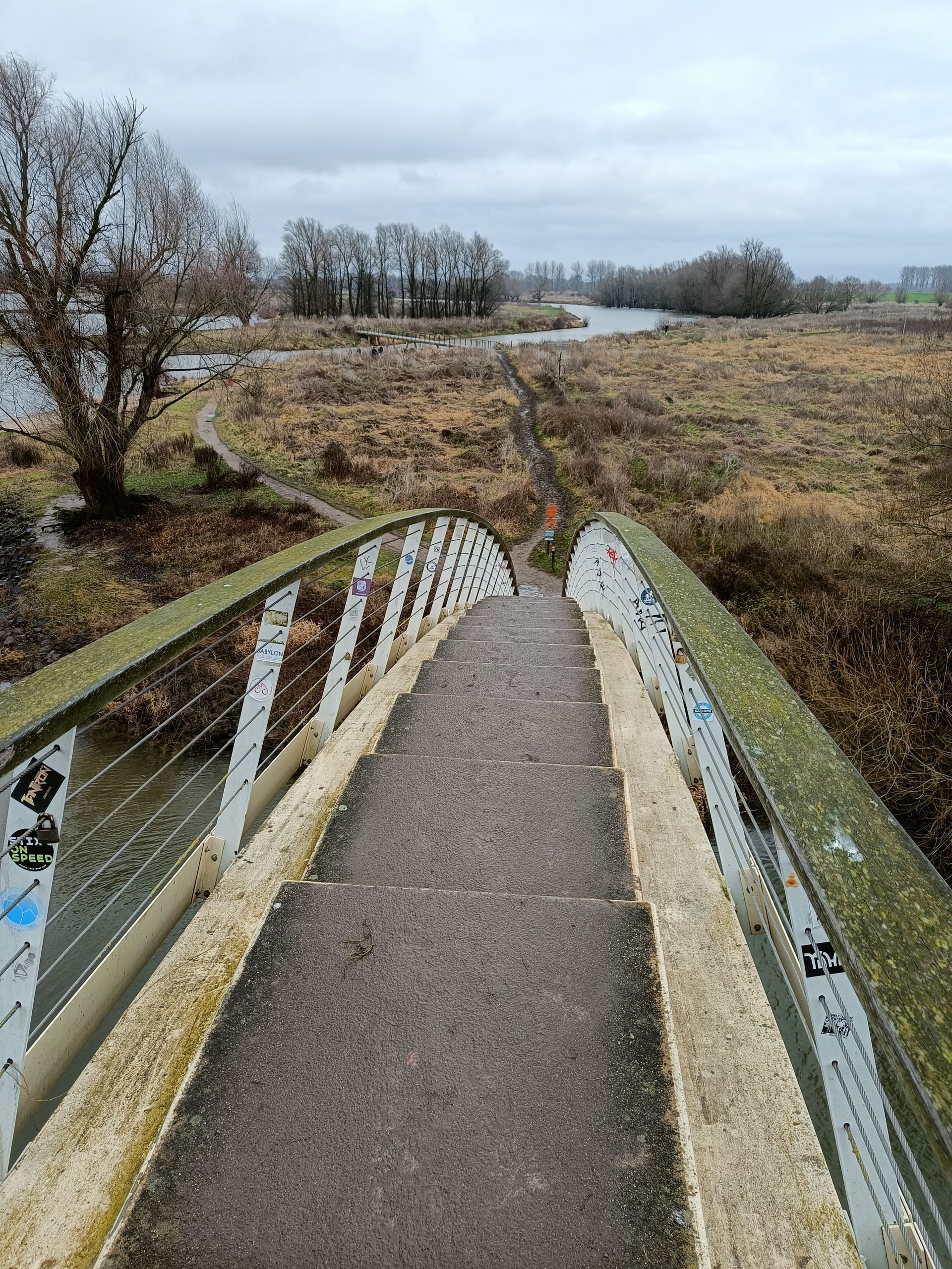Een brug overspant een rivier en leidt naar een pad door een grasrijk landschap onder een bewolkte hemel.