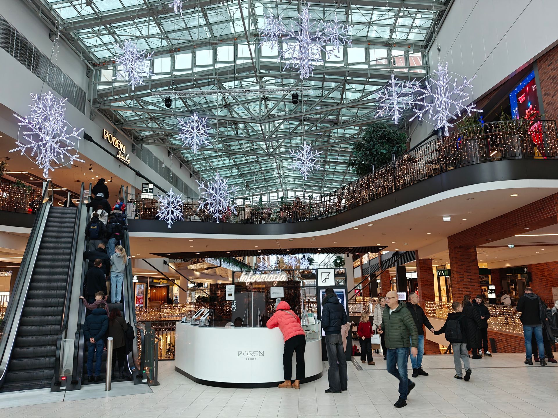 Interieur van een winkelcentrum, mensen die langs een winkelbalie lopen, roltrappen en hangende decoratieve sneeuwvlokken.