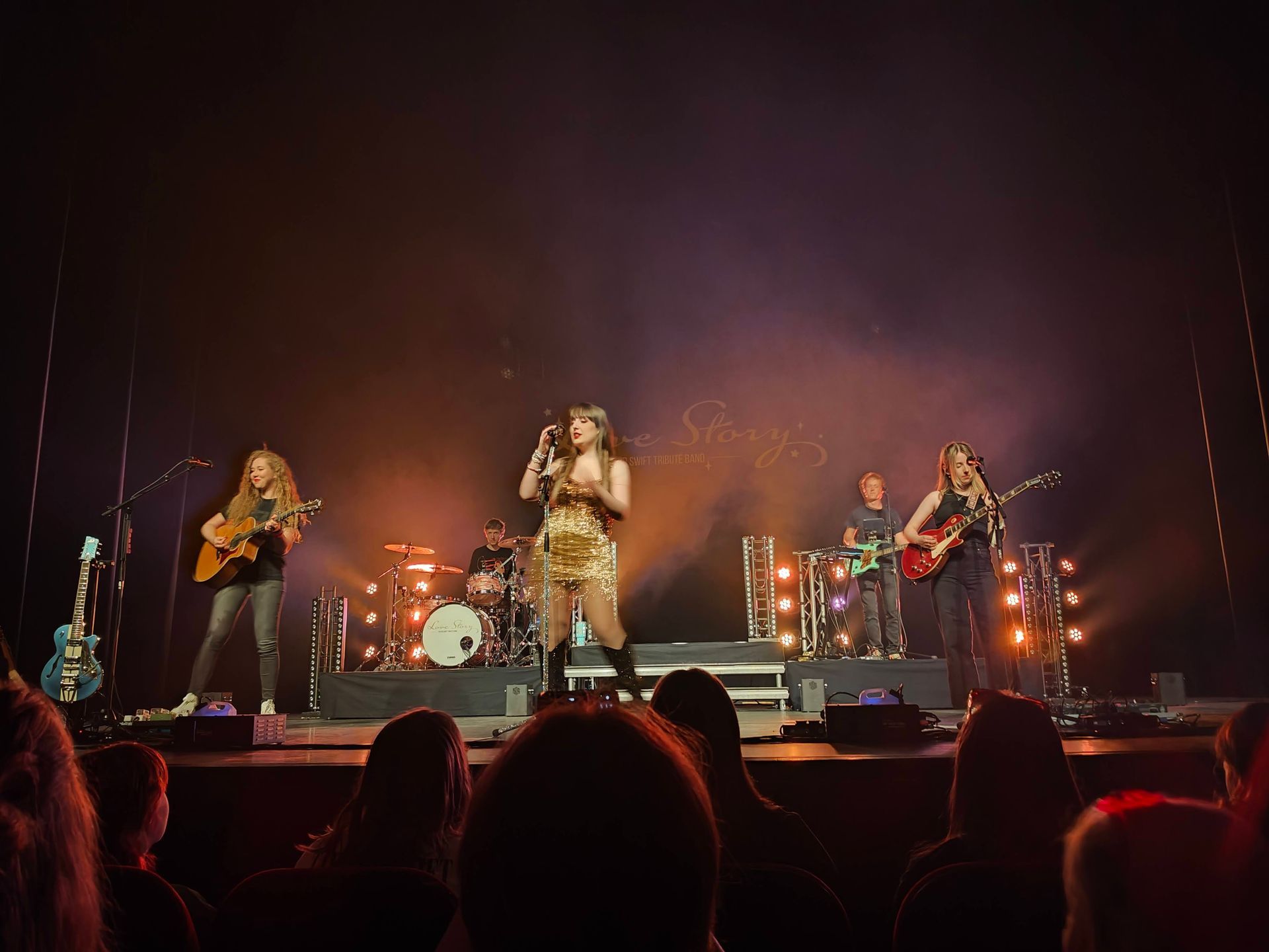 Band treedt live op op een schemerig verlicht podium met drums, gitaren en warme oranje verlichting.