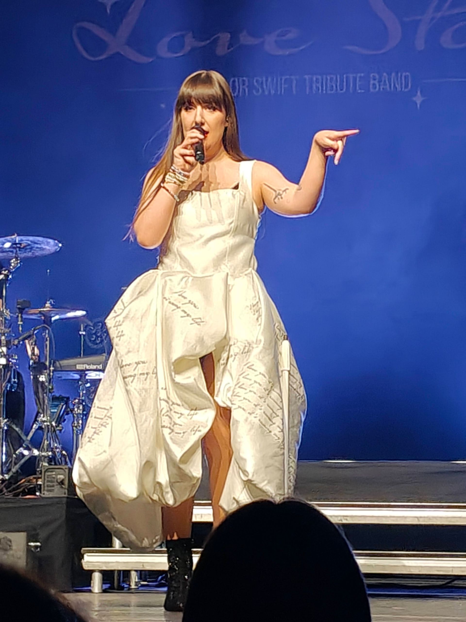 Een zangeres in een witte jurk treedt op het podium op met een microfoon, met een blauw concertdecor achter haar.