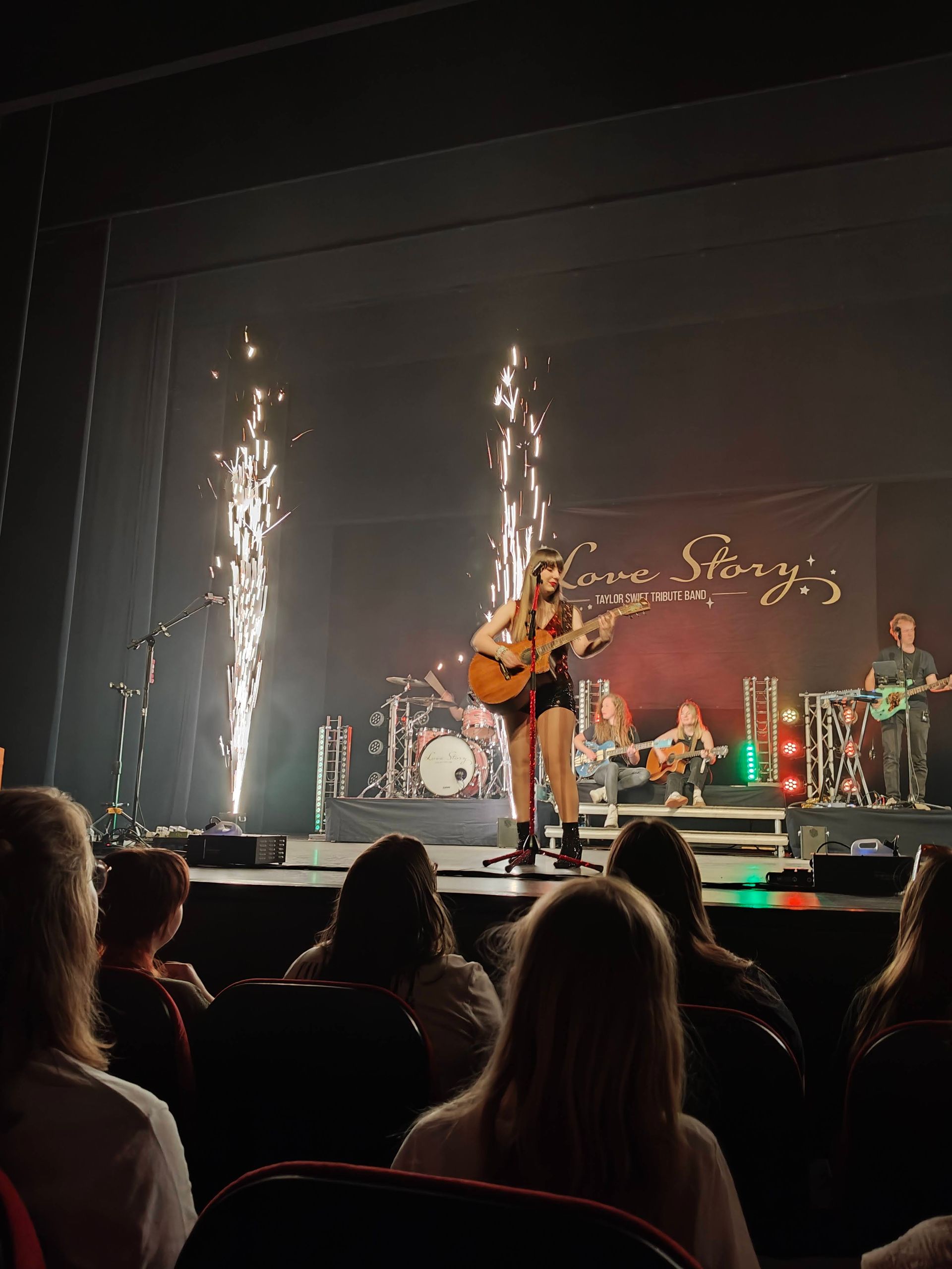 Zangeres treedt op het podium op met gitaar, vuurwerk en een zittend publiek op de voorgrond.