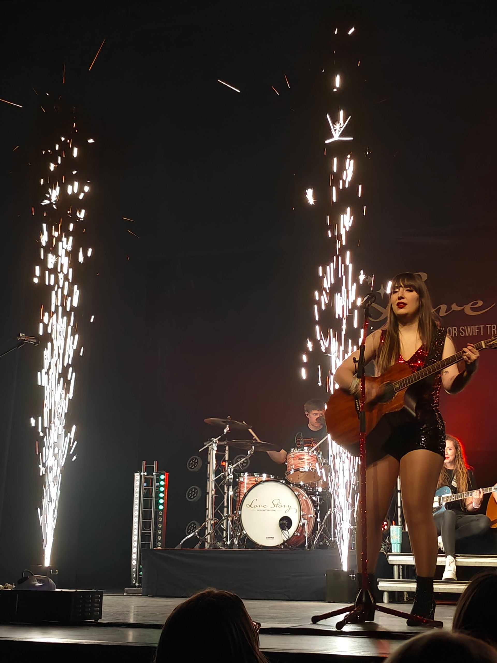 Een zangeres met gitaar staat op het podium en zingt te midden van heldere fonteinen en een drumstel achter haar.