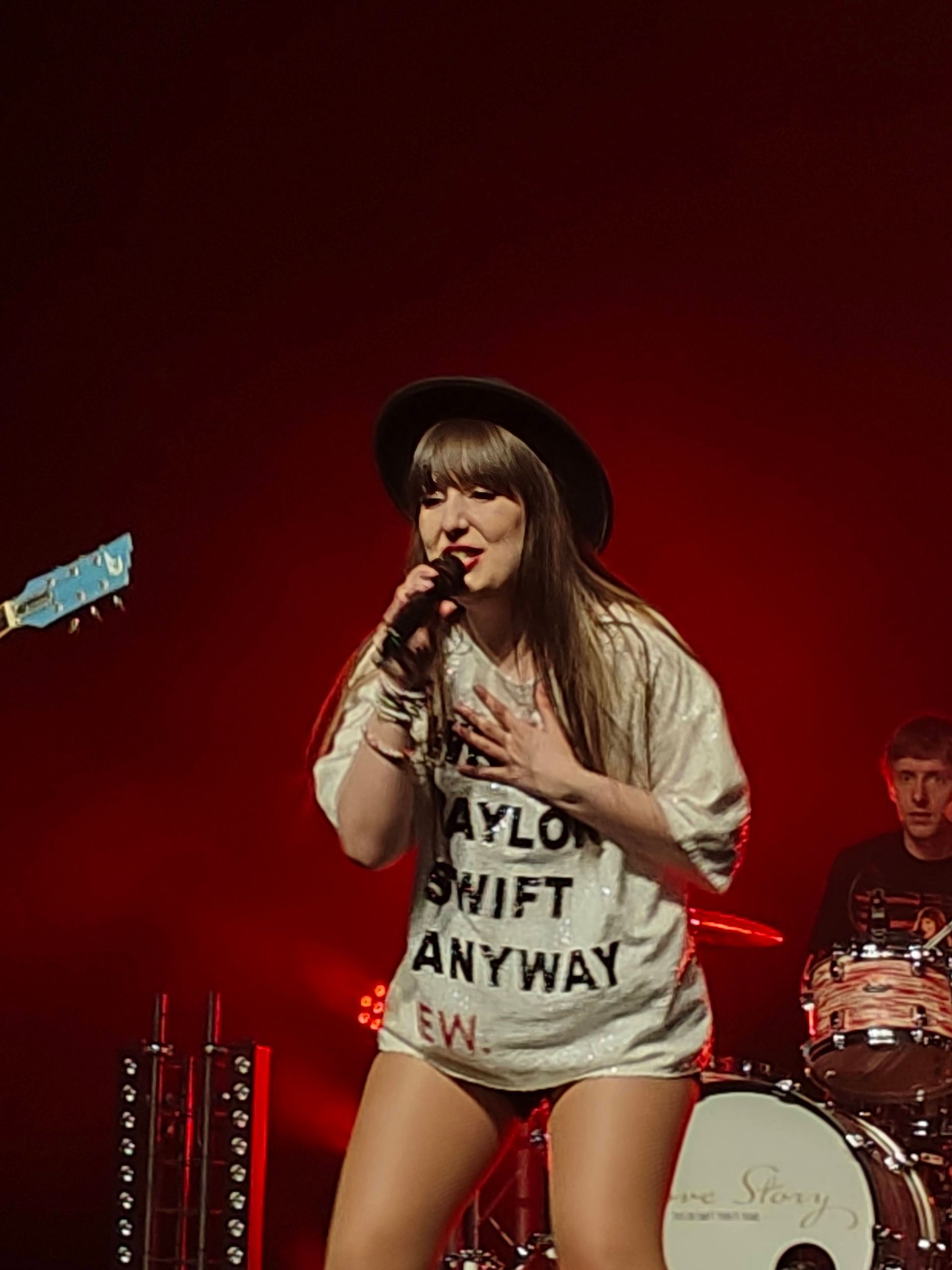 Zangeres treedt op het podium op met een microfoon, draagt ​​een hoed en een wit Taylor Swift-shirt tegen een rode achtergrond.