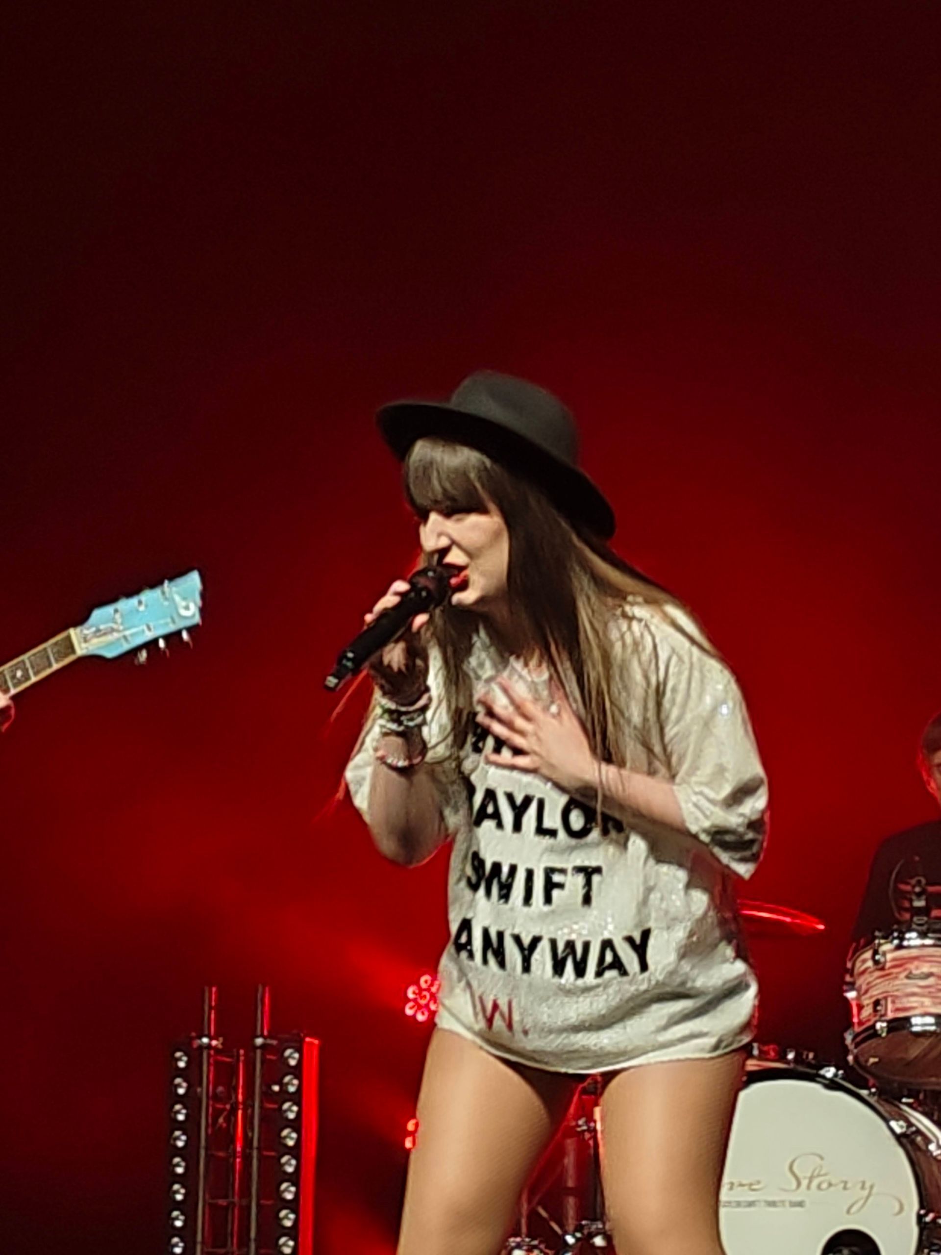 Zangeres met zwarte hoed en wit T-shirt zingt op het podium onder rode lichten.
