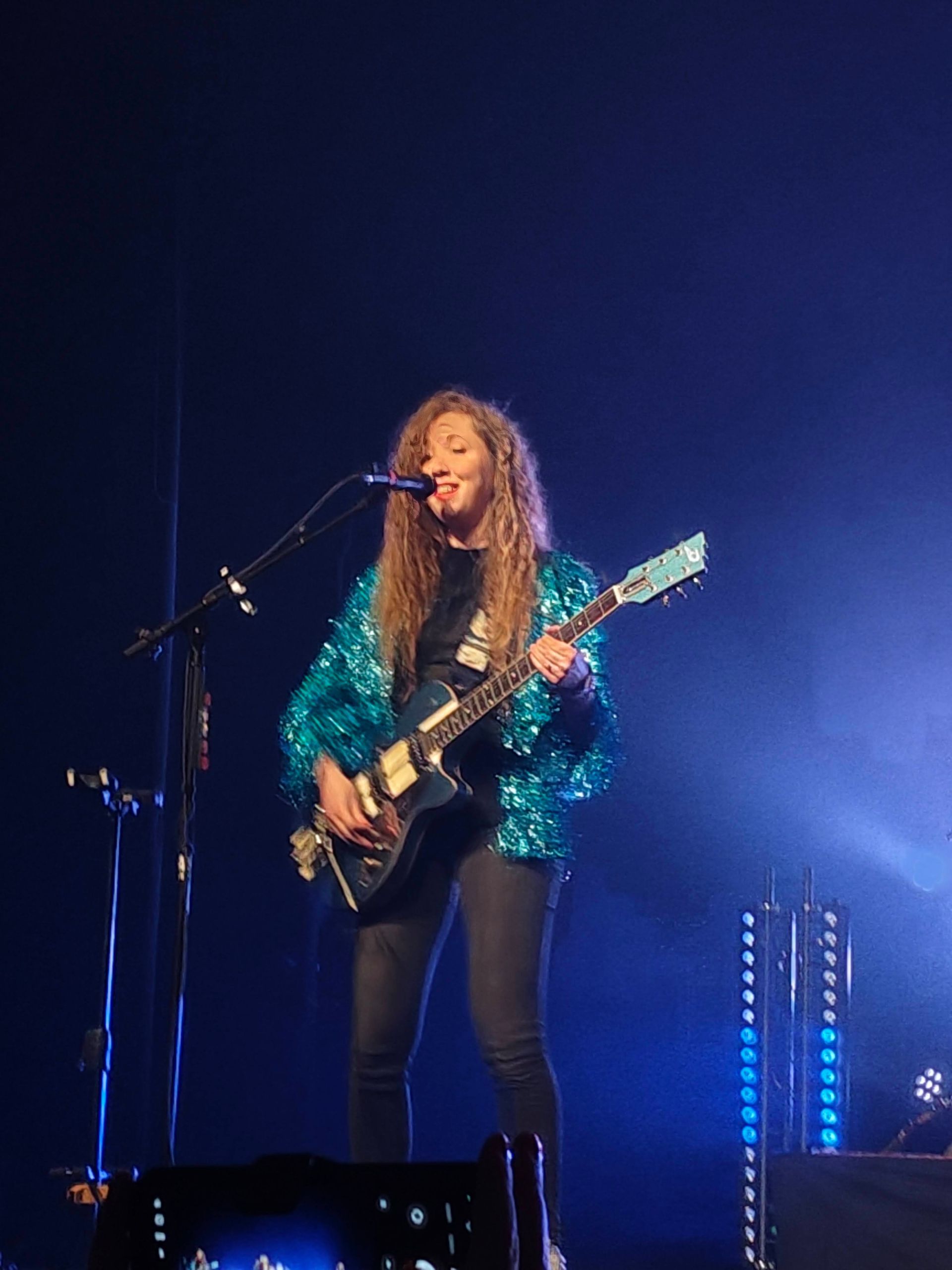 Zangeres treedt op het podium op met een elektrische gitaar, gekleed in een turquoise jasje onder blauwe verlichting.