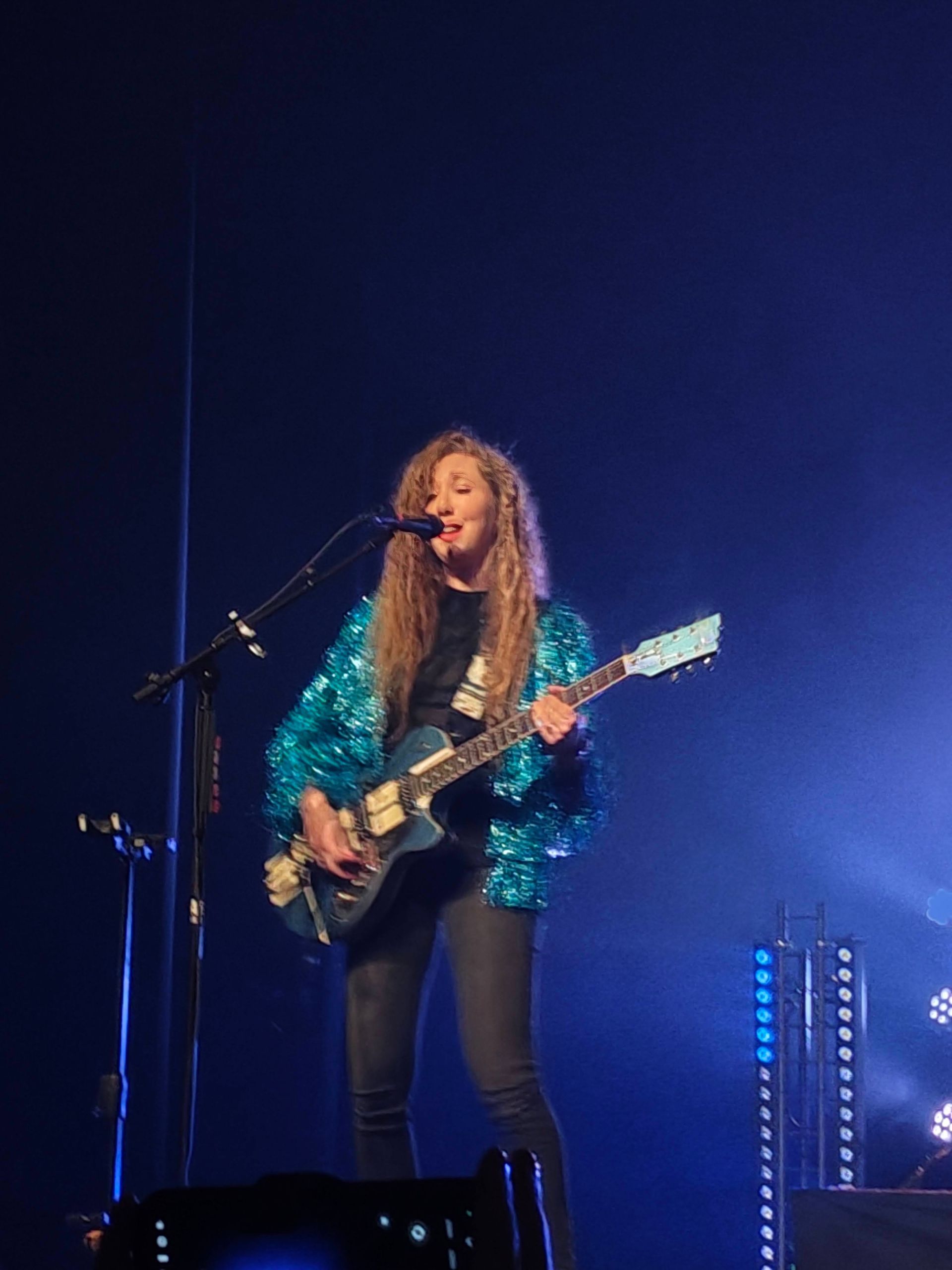 Zangeres in een turquoise jasje treedt op met een elektrische gitaar op het podium onder blauwe lichten.