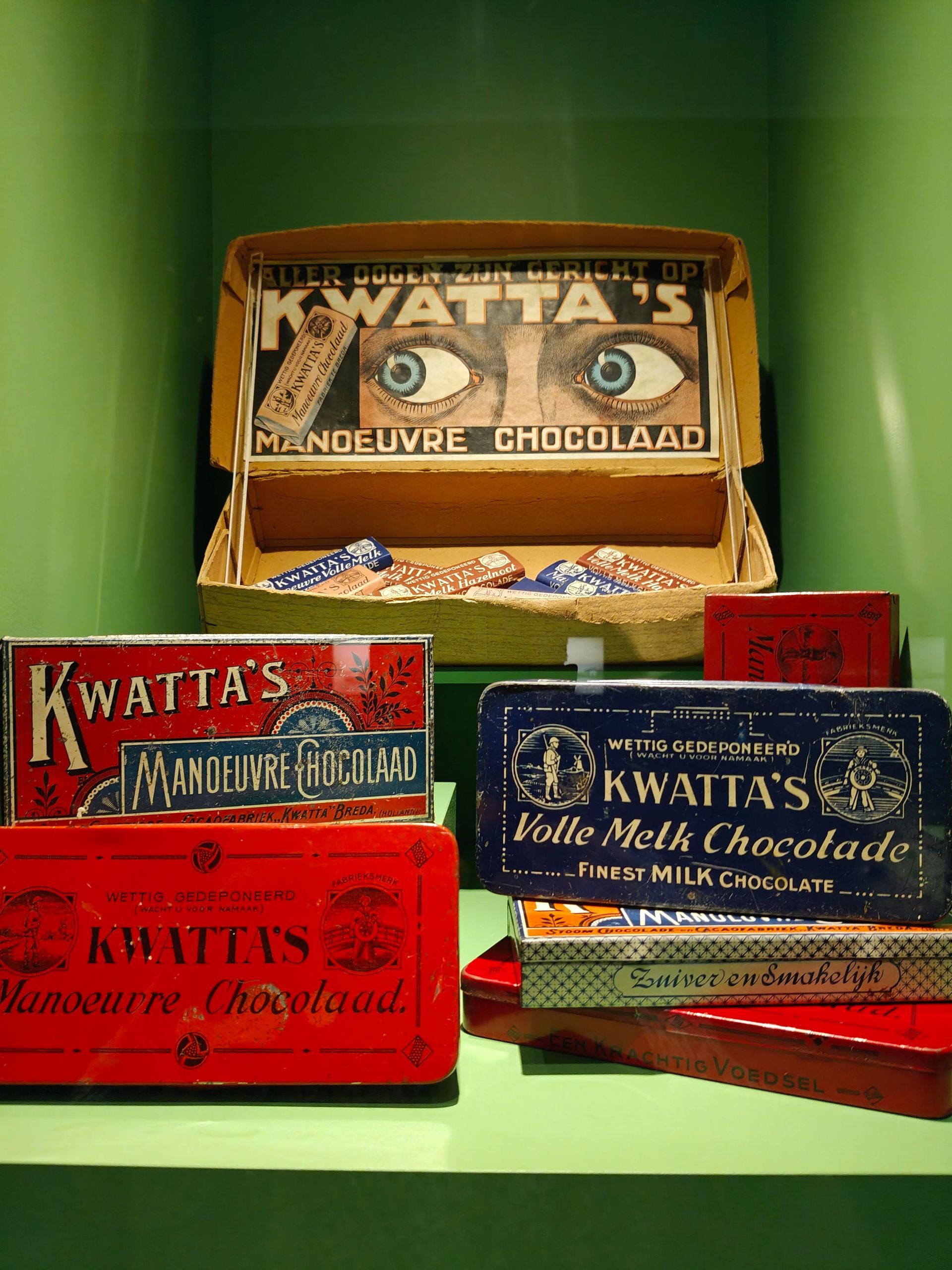 Diverse vintage Kwatta-chocoladeblikken en -dozen opgestapeld op een groene achtergrond.